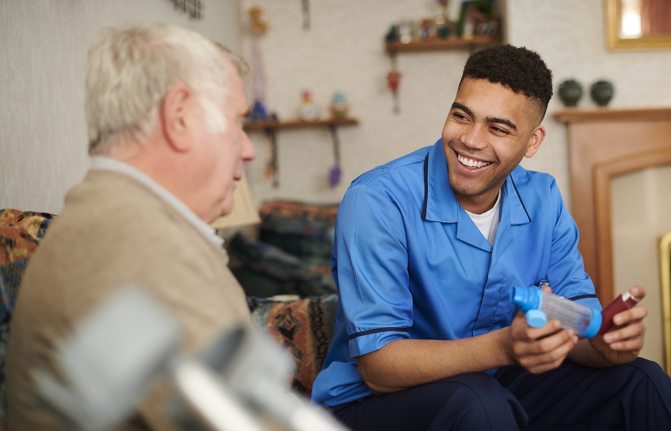 A young support worker talking to an older man in his home. He is also assisting the man to take his inhaler.