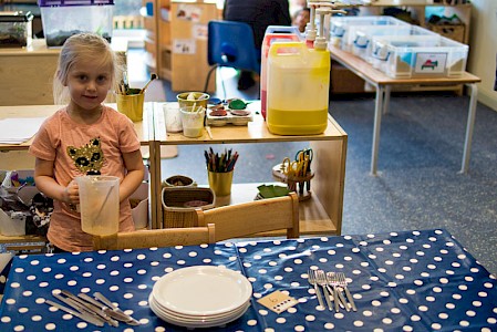 A young girl is setting the table in the nursery. She uses a card with six dots to help her count the right number of forks and knives.
