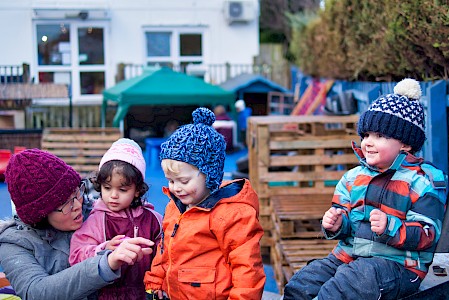 A childcare centre’s garden. An early years practitioner is engaging with three young children in winter clothing.